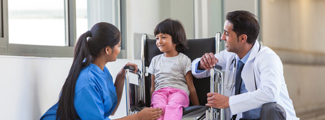 Una enfermera y un médico hablan con una niña sentada en una silla de ruedas