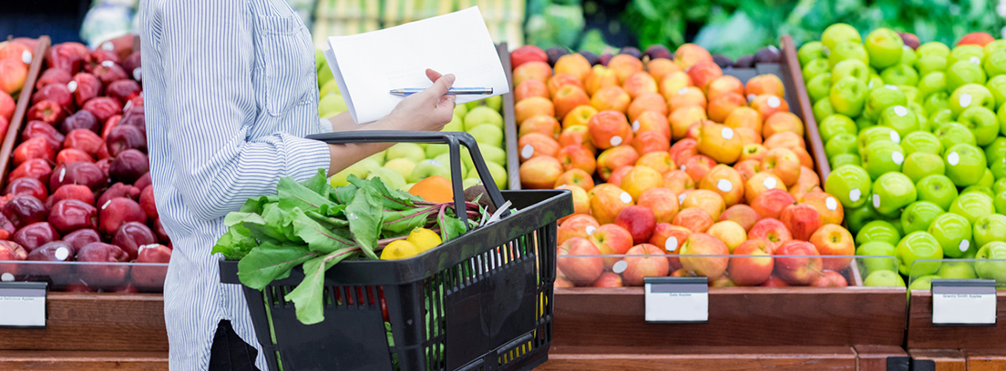 Mujer en primer plano con una cesta llena de verduras en la zona de frutas de un supermercado