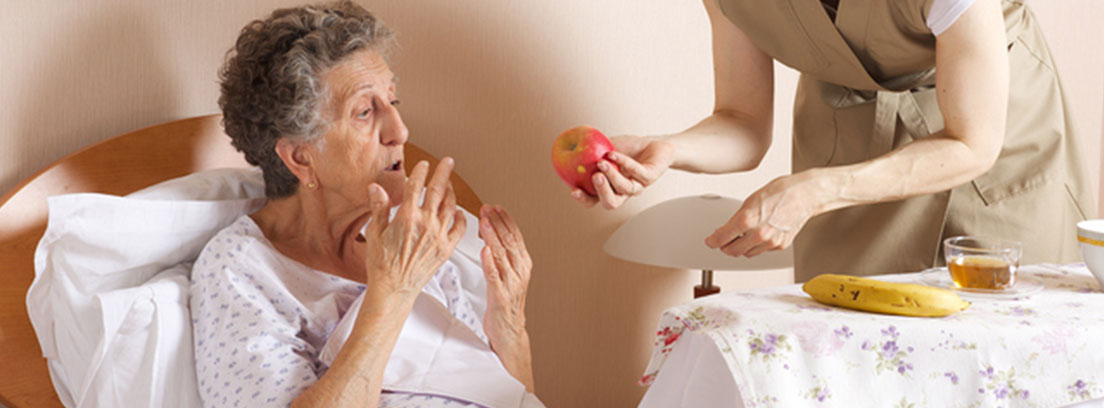 Una cuidadora ofrece una manzana a una anciana que se encuentra en la cama