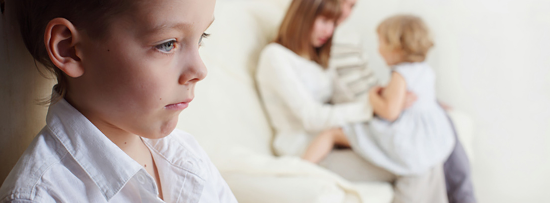 Niño triste con la mirada perdida mientras su madre juega con una hermana pequeña