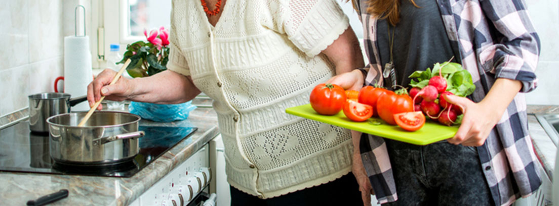 Una mujer mayor cocina y otra joven le acerca unos tomates