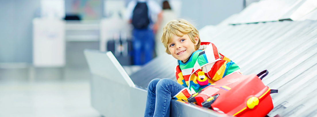 Un niño sentado en la cinta de recogida de maletas de un aeropuerto junto a su maleta roja