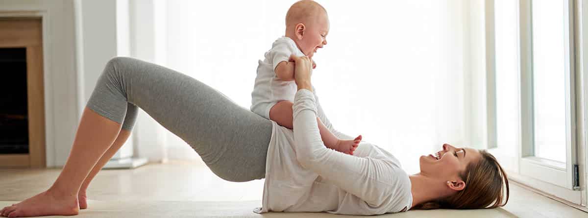 Mujer tumbada en una colchoneta realizando ejercicios con el bebé sobre su abdomen