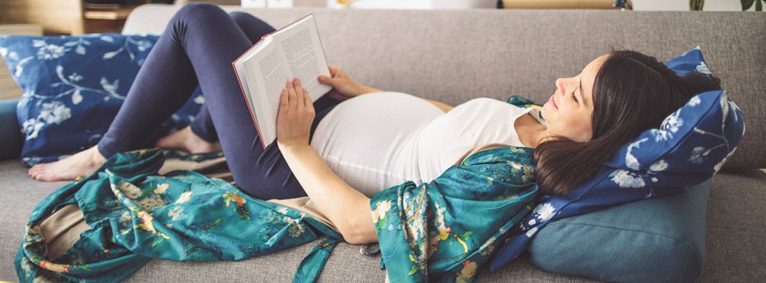 Cuidados en el primer trimestre de embarazo: mujer embarazada tumbada en el sofá leyendo un libro