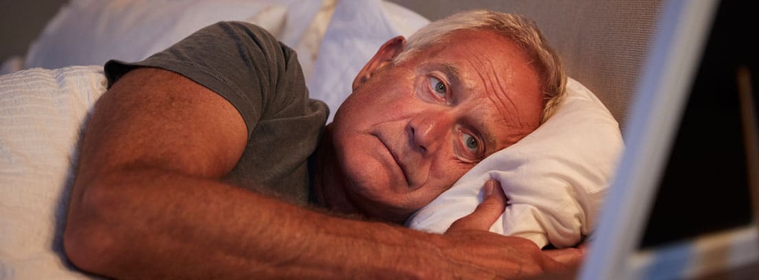 Hombre mayor tumbado en la cama mirando el marco de la foto e intentando concililar el sueño