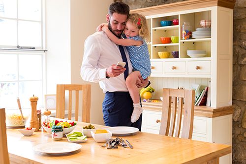 padre con su hija en brazos mirando el móvil al lado de una mesa con desayunos