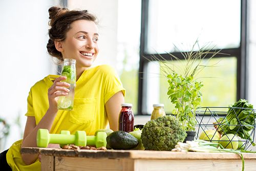 mujer joven bebiendo agua con limón, mesa de madera con unas mancuernas y variedad de verduras