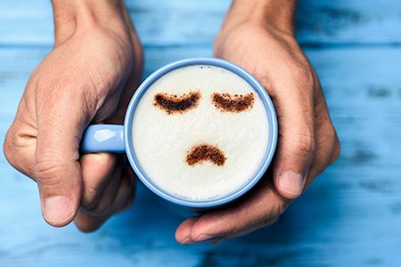 manos sujetando una taza de café y dibujado en la espuma una carita triste