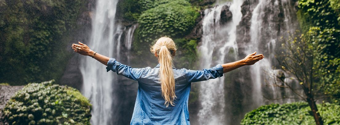 mujer rubia de espaldas contemplando una cascada en el bosque