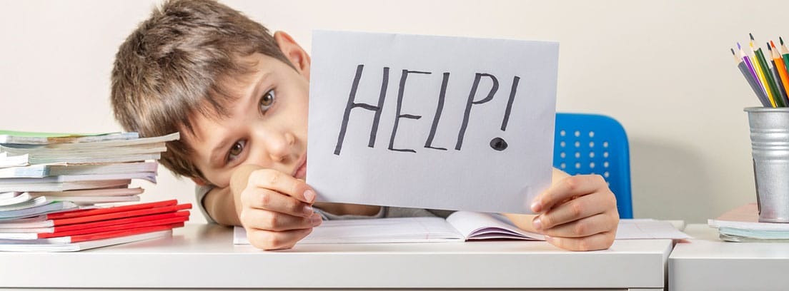 Problemas de aprendizaje en niños: niño medio tumbado sobre una mesa de estudio con una hoja en la mano con la palabra ayuda