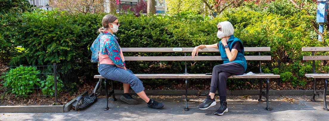 ¿Cómo afecta la nueva normalidad a nuestra salud? : dos mujeres sentadas en un banco de la calle con mascarillas y guardándo la distancia de seguridad