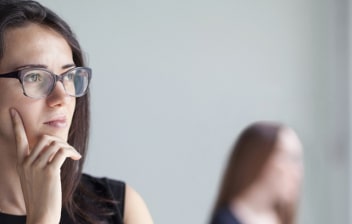 ¿Qué es el trastorno de personalidad evitativa?: chica joven con gafas y las manos en la barbilla dudando, al fondo dos mujeres hablando