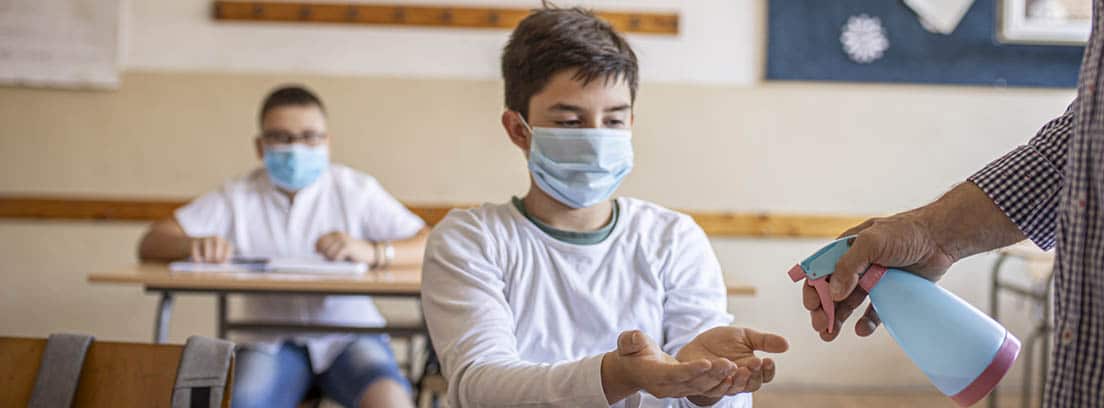 Vuelta al cole y COVID-19: niños con mascarilla en un aula y profesor echando gel en las manos