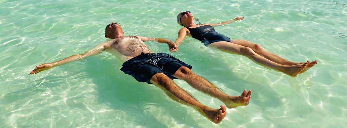 Beneficios del agua del mar para la piel: Pareja madura tomados de la mano mientras flota en las aguas poco profundas de la playa del Mar Muerto