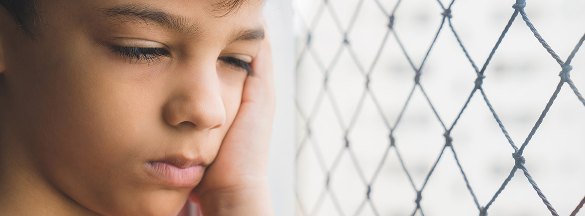 Niño al lado de una ventana con los ojos cerrados y la mano apoyada en un lado de la cara