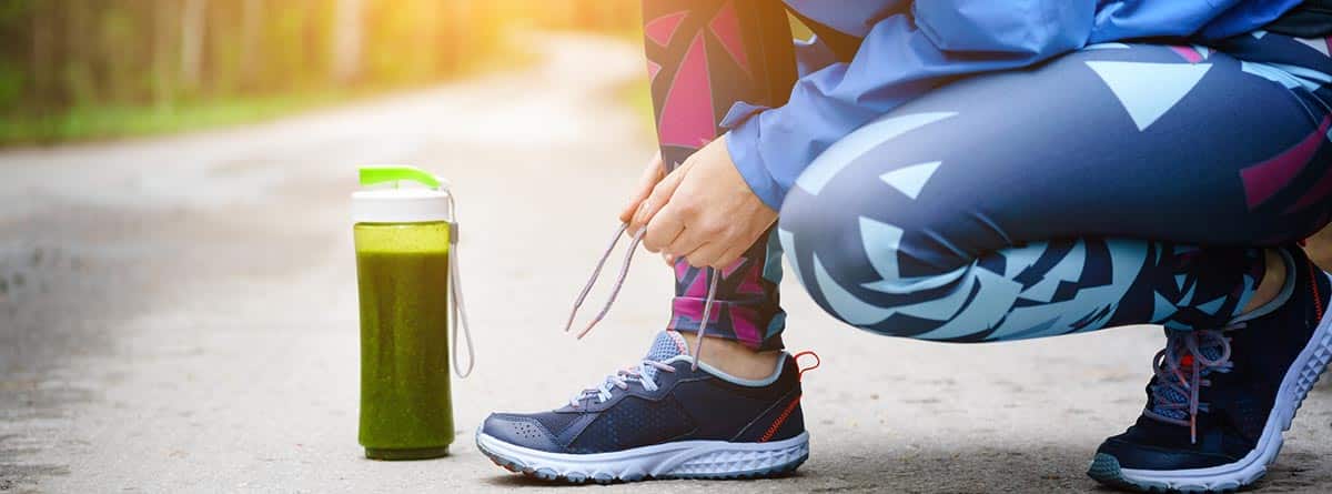 Suplementos deportivos, ventajas y desventajas: corredor femenino atándose los cordones de la zapatilla y un batido de color verde al lado
