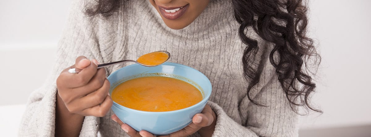 Mujer vistiendo un suéter de invierno, sentada en la mesa de la cocina comiendo una sopa caliente