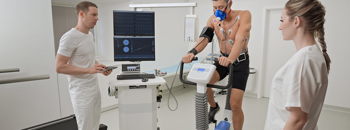 Hombre maduro haciendo una prueba de esfuerzo cardiopulmonar en bicicleta en una clínica supervisada por médicos masculinos y femeninos.