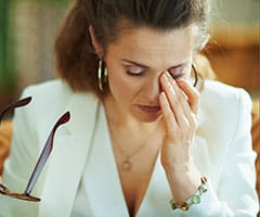 ¿Qué es el Síndrome de Sjögren?: mujer cansada de mediana edad con blusa blanca y chaqueta con gafas frotándose los ojos en la sala de estar