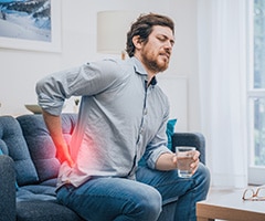Causas del dolor de riñones: hombre sentado en un sofá con la mano apoyada en la zona de los riñones en color rojo