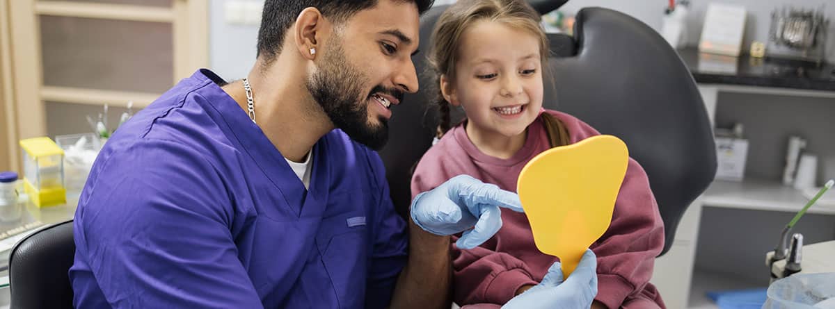 Niña disfrutando de su sonrisa con dientes mirando el espejo mientras un joven dentista lo sostiene.