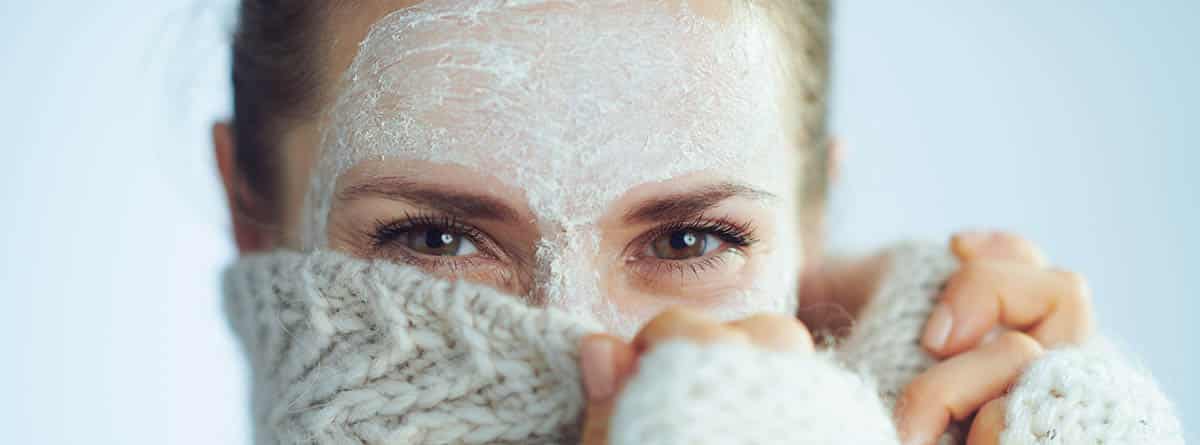 Recomendaciones para tener una piel perfecta en invierno: mujer tapándose medio rostro con ropa de invierno y crema en la cara