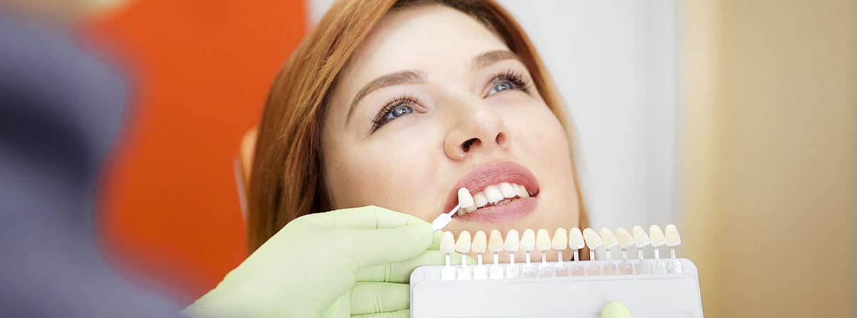 Coronas dentales y tipos: mujer en la consulta del odontólogo comprobando el color del esmalte del diente para futura corona dental