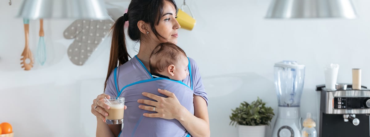 Portear a un bebé, ¿es realmente bueno?: madre muy joven con un bebé en un cabestrillo bebiendo café en la cocina de casa.