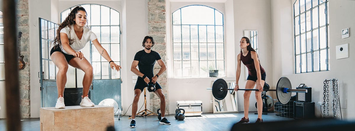 Por qué hacer ejercicios de fuerza y resistencia: dos mujeres y un hombre en un gimnasio realizando diferentes tipos de ejercicio de fuerza