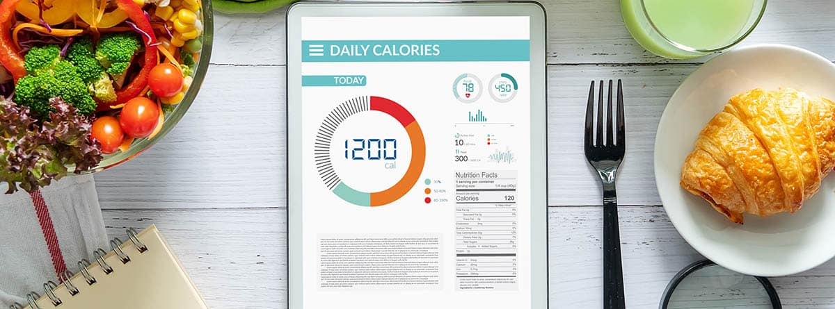 Déficit calórico: tableta con aplicación de contador de calorías en la pantalla en la mesa de comedor con ensalada, jugo de fruta, pan y vegetales