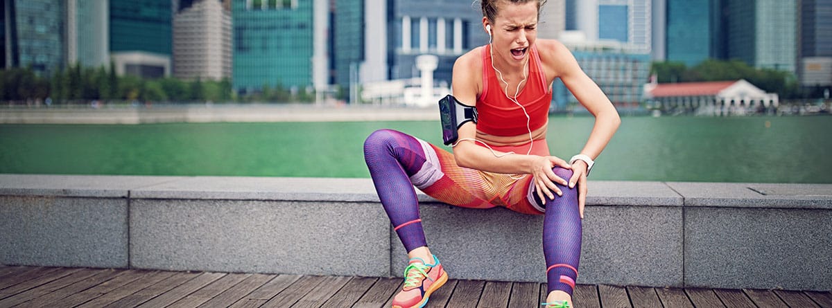 Cómo prevenir los calambres al hacer deporte: chica deportista sentada en la calle con mucho dolor en la pierna