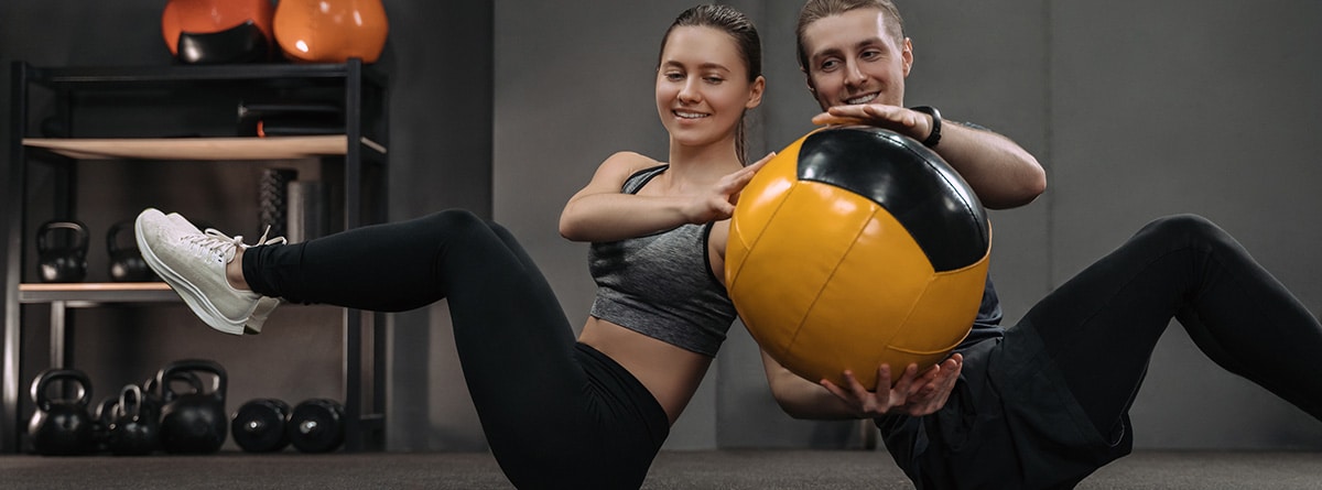 Entrenamiento HIIT: Pareja joven sana haciendo ejercicios de abdominales con balón medicinal en el gimnasio oscuro