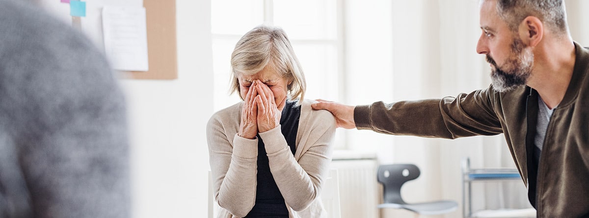 ¿Como actuar ante una crisis de ansiedad?: Una mujer mayor deprimida llorando durante una terapia de grupo, otras personas consolándola.