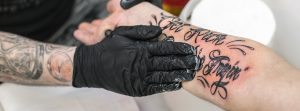 ¿Cómo curar un tatuaje infectado?: brazo tatuado un poco rojo y manos con guantes negros aplicando una pomada