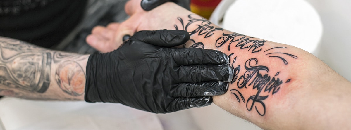 ¿Cómo curar un tatuaje infectado?: brazo tatuado un poco rojo y manos con guantes negros aplicando una pomada