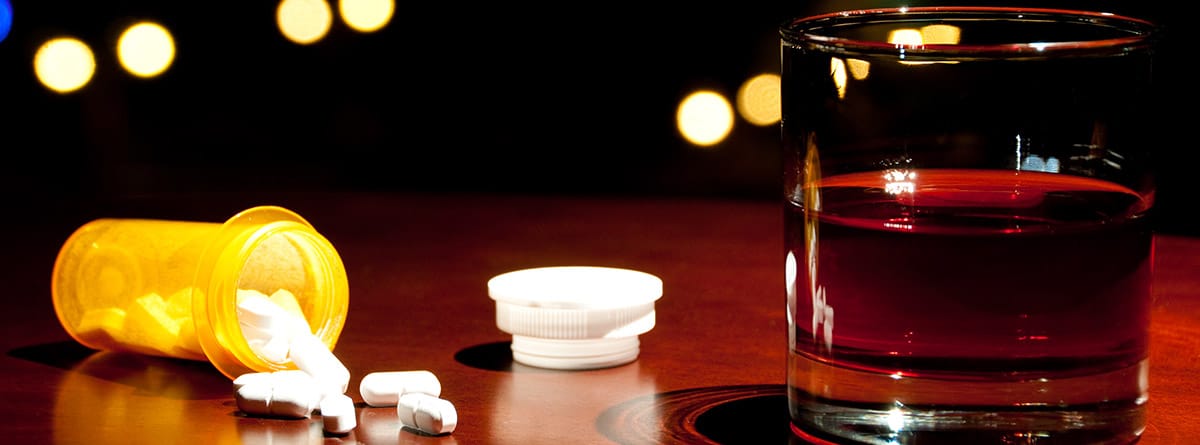 Vaso con bebida alcohólica junto a una botella de pastillas derramada sobre una mesa de madera, con las luces de la ciudad al fondo.