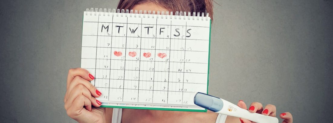 Mujer joven escondida detrás de un calendario menstrual y mostrando una prueba de embarazo positiva