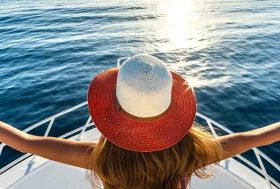 Mujer de espaldas con sombrero puesto en la parte delantera de un barco con el mar de fondo.