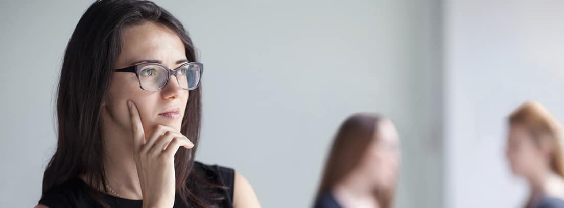 Evitar dificultades ¿Qué es el trastorno de personalidad evitativa?: chica joven con gafas y las manos en la barbilla dudando, al fondo dos mujeres hablando