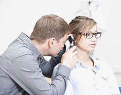 Otitis, infección de oídos: Un doctor masculino examinando el oído de una paciente con un otoscopio.