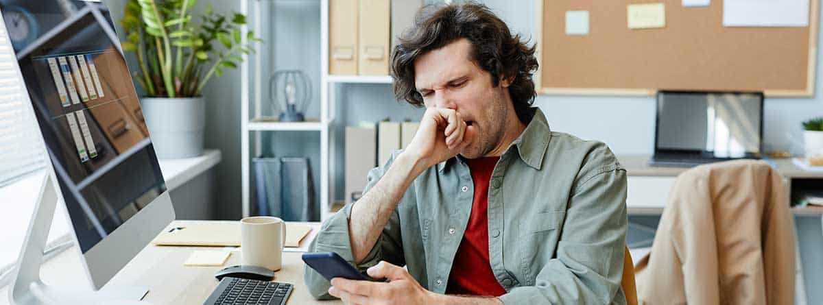 procrastinación1 Cómo vencer a la procrastinación: un joven caucásico bostezando en el lugar de trabajo de oficina y navegando por las redes sociales por teléfono